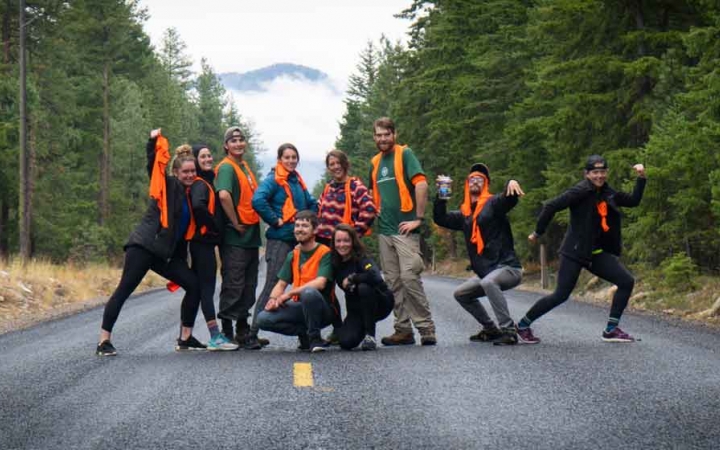 a group of gap year students wearing orange vests pause for a photo on a road between green trees. They have been doing service projects with outward bound. 